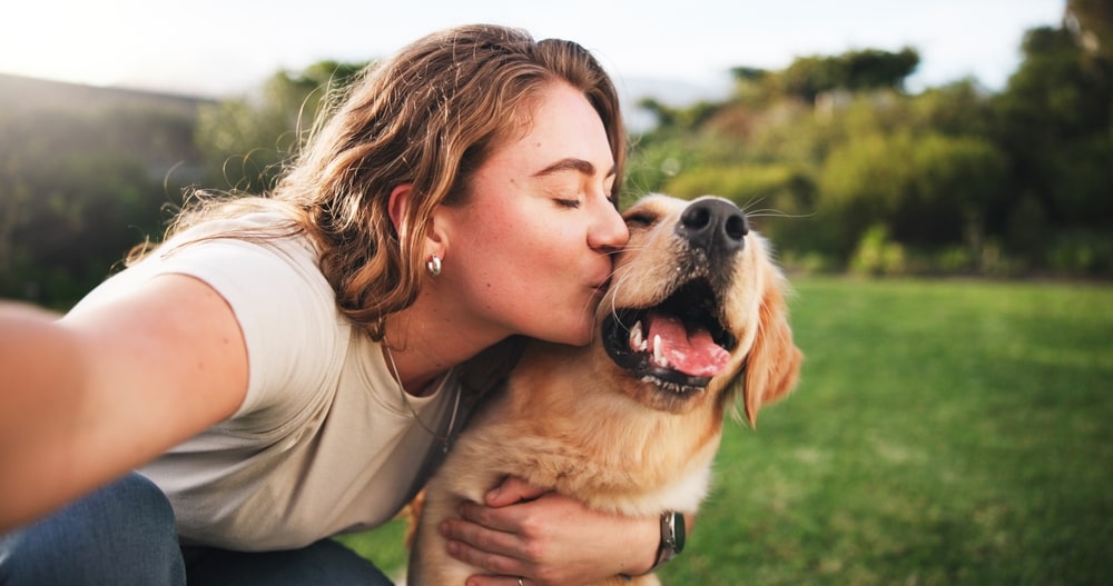 Pet parentkissing her happy Golden Retriever outdoors on a sunny day.