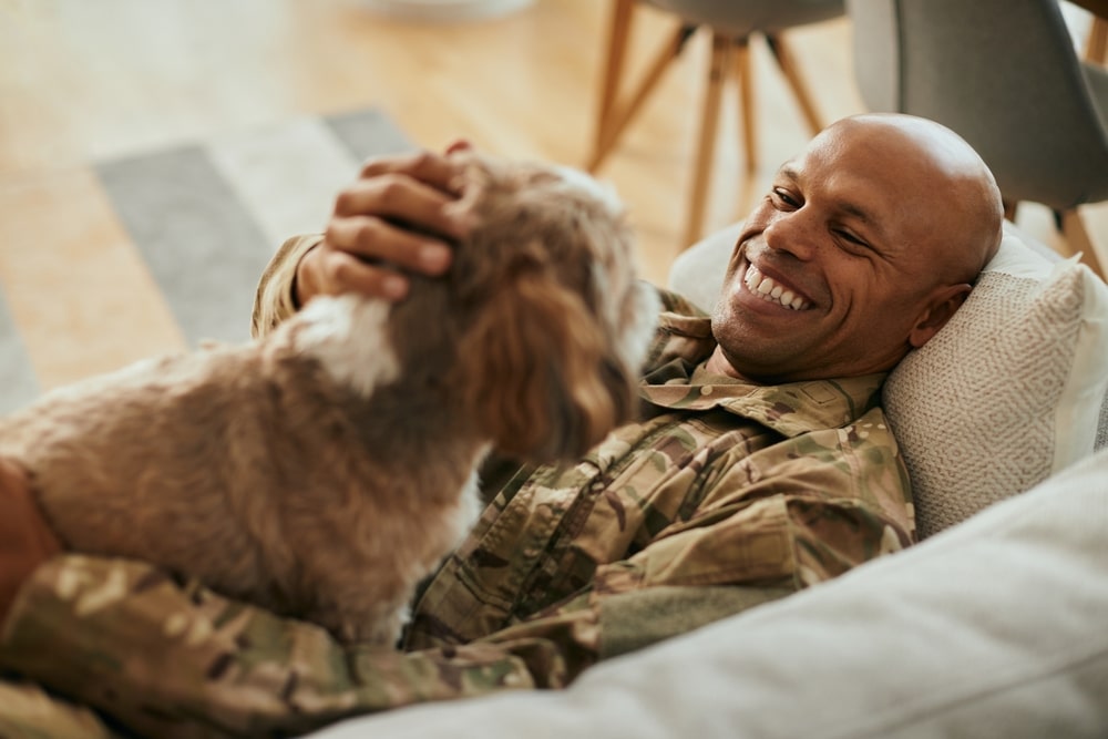 Young happy African American veteran cuddling his dog while relaxing on the sofa.
