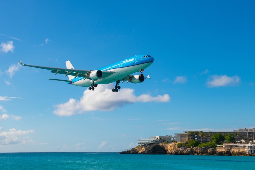 KLM airplane flying low over turquoise ocean, approaching a coastal runway on a clear blue-sky day.